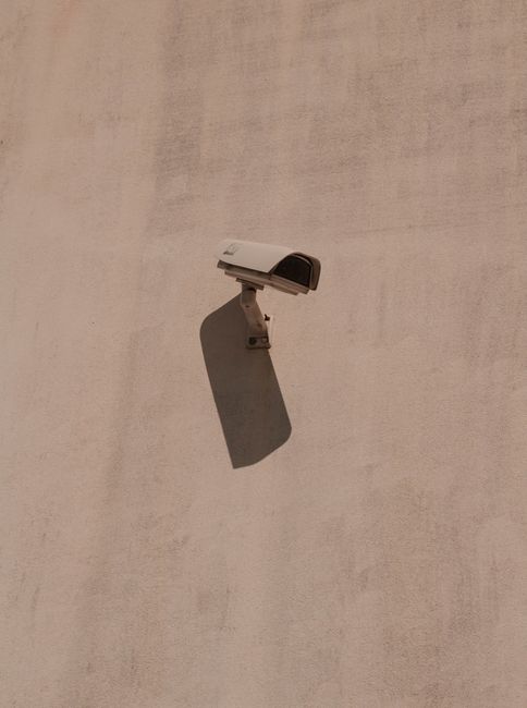 A minimalist photograph of a security camera casting a shadow on a plain concrete wall.